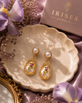 A pair of gold dangle earrings with white pearl-like round studs and teardrop-shaped clear resin pendants containing pink and green floral details, displayed on a scalloped beige dish with purple flowers, satin fabric, and an Irises Jewels box in the background.