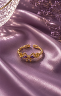 Gold adjustable bow ring with lavender crystal stones displayed on soft lavender satin fabric.