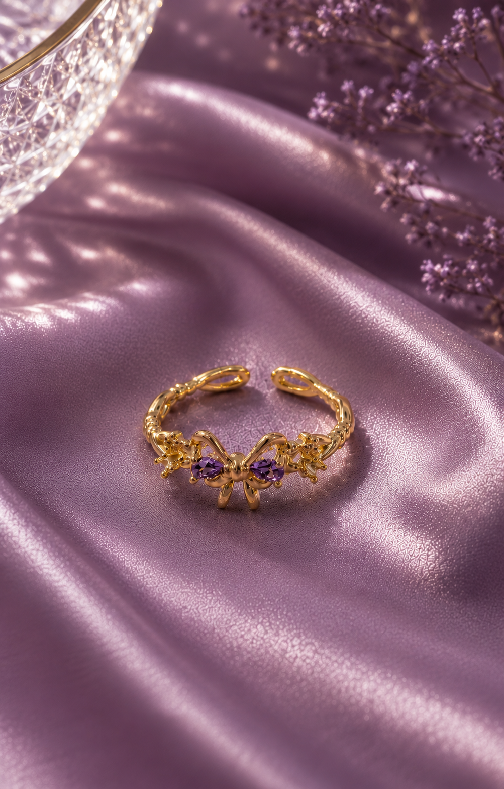 Gold adjustable bow ring with lavender crystal stones displayed on soft lavender satin fabric.