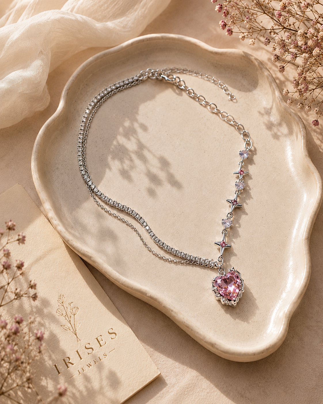 A silver-toned chain necklace with a heart-shaped pink gemstone pendant, displayed on a beige ceramic tray with dried flowers and soft fabric around it. The brand name 'IRISES JEWELRY' is visible on a card.