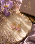 A pair of gold heart-shaped drop earrings with sparkling pink stones, displayed on a scalloped cream tray with purple flowers, lavender silk, and a box labeled Irides Jewels.