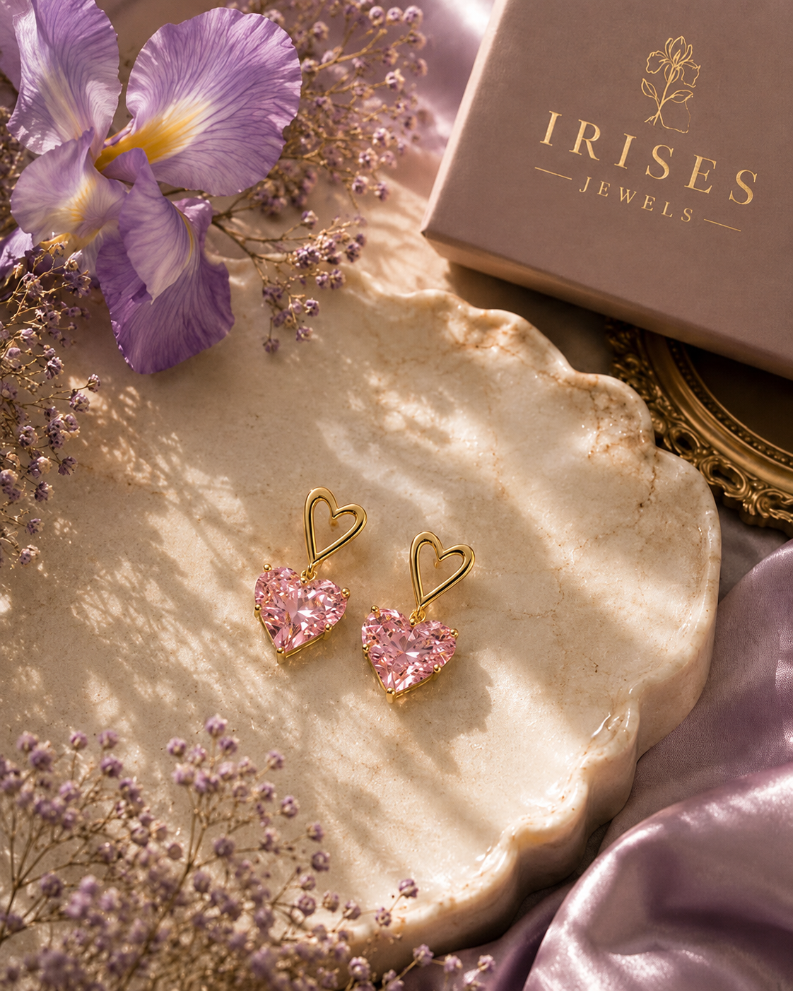 A pair of gold heart-shaped drop earrings with sparkling pink stones, displayed on a scalloped cream tray with purple flowers, lavender silk, and a box labeled Irides Jewels.