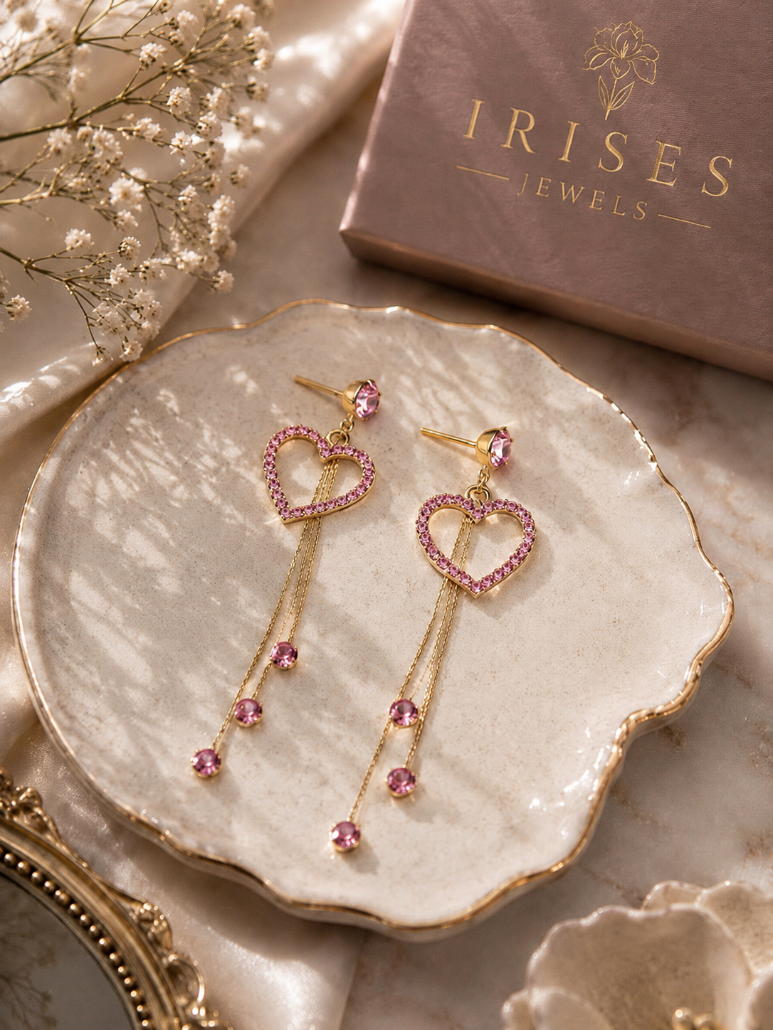 Blush Heart Tassel Drop Earrings featuring sparkling pink crystal heart frames with elegant gold tassel chains. Pair of gold dangle earrings with heart-shaped crystal borders and pink gemstone drops, displayed on a scalloped cream dish with a pink 'IRISES JEWELS' box in the background.