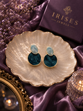 Ocean Mist Marble Drop Earrings featuring elegant blue marbled resin discs with a premium gold-tone finish and oval light blue stones on top and round dark teal-blue stones below, displayed on a scalloped cream dish with purple satin fabric.