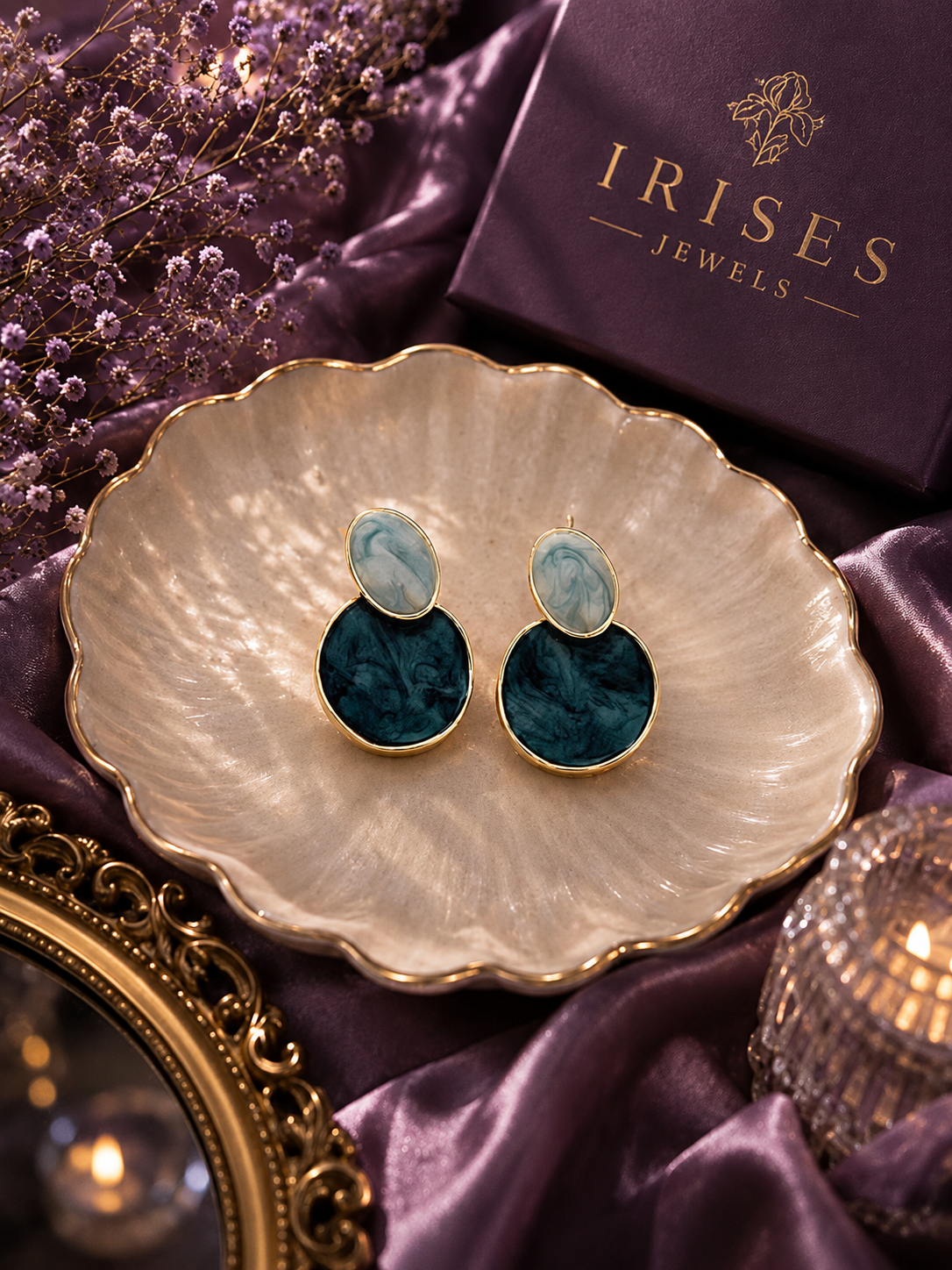 Ocean Mist Marble Drop Earrings featuring elegant blue marbled resin discs with a premium gold-tone finish and oval light blue stones on top and round dark teal-blue stones below, displayed on a scalloped cream dish with purple satin fabric.