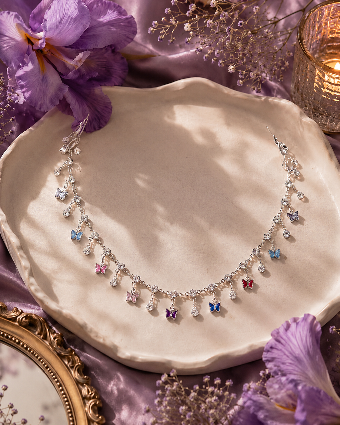 A delicate silver-toned necklace with small clear stones and colorful butterfly-shaped charms in pink, blue, purple, and red, displayed on a cream ceramic dish with purple flowers and purple satin fabric.