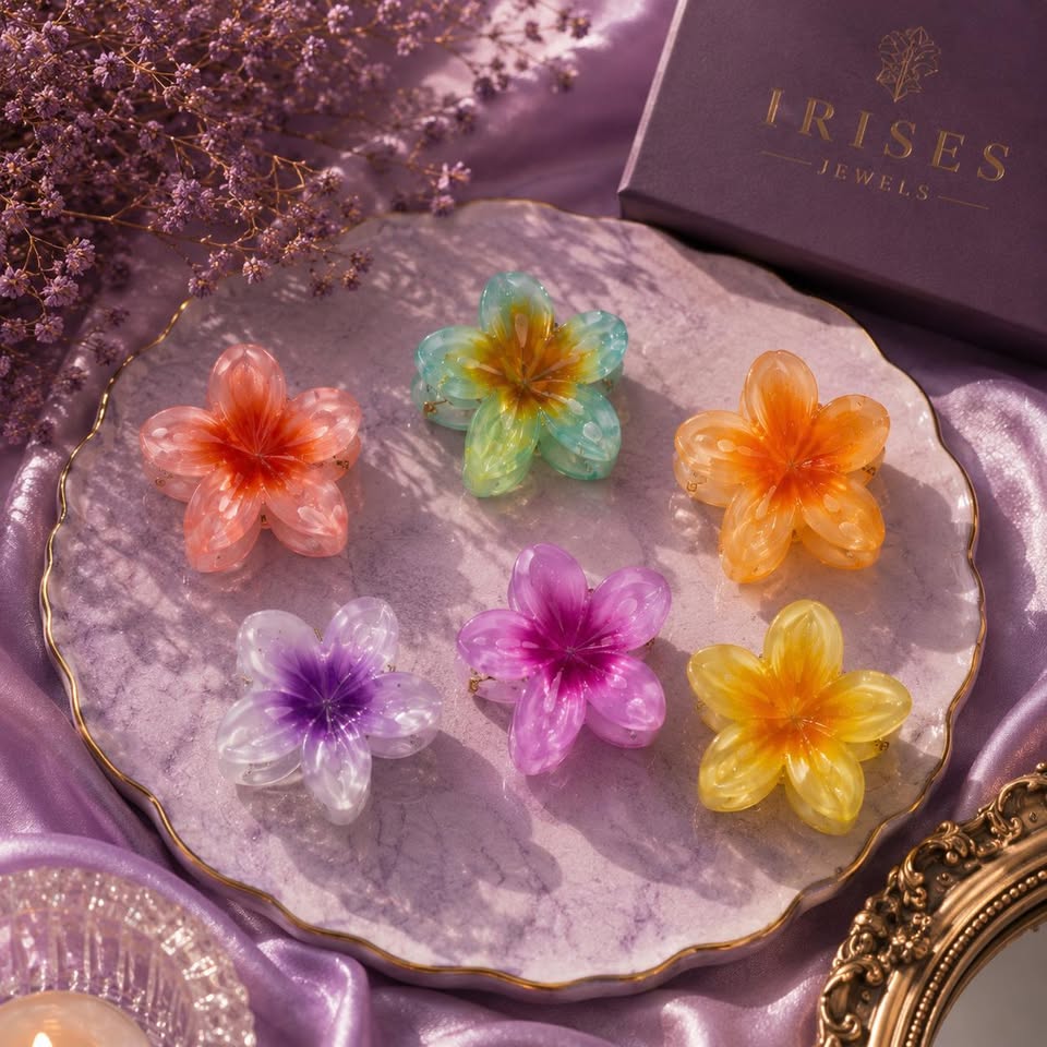 Five translucent flower-shaped jewelry pieces in peach, mint, orange, purple, and yellow are arranged on a scalloped lavender tray, with a purple Irides Jewels box and decorative dried flowers in the background.