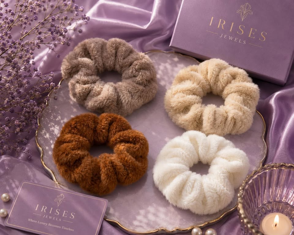 Four fluffy faux-fur hair scrunchies in taupe, cream, brown, and off-white, arranged on a decorative tray with purple satin fabric, a purple Irises Jewelry box, a card, and a lit candle.