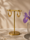 Textured gold hoop earrings displayed on a gold jewelry stand with soft beige background and purple iris flower styling.
