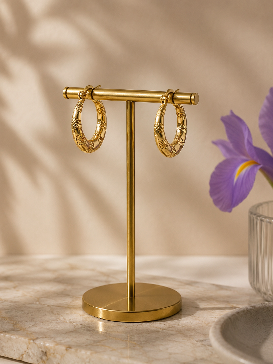 Textured gold hoop earrings displayed on a gold jewelry stand with soft beige background and purple iris flower styling.