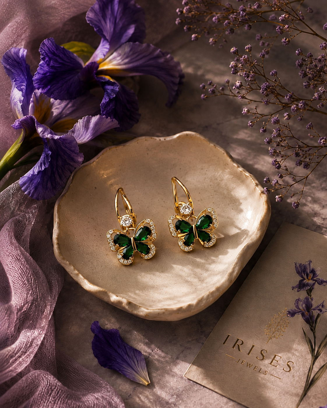 Gold butterfly drop earrings with green enamel centers and clear crystal accents, displayed on a textured beige dish with purple flowers and a card reading Trisles Jewellery.