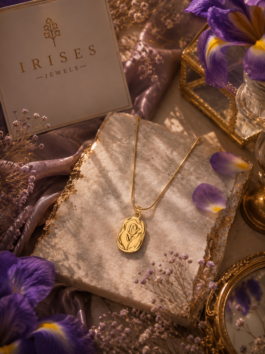 A gold pendant necklace with an engraved rose design on a delicate chain, displayed on a textured stone slab. The setting features purple irises, dried flowers, purple silk, and a gold-framed mirror, with a branded 'IRISES JEWELLERY' box visible in the background.