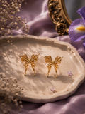 Gold butterfly stud earrings displayed on a ceramic tray with lavender flowers and soft silk fabric styling.