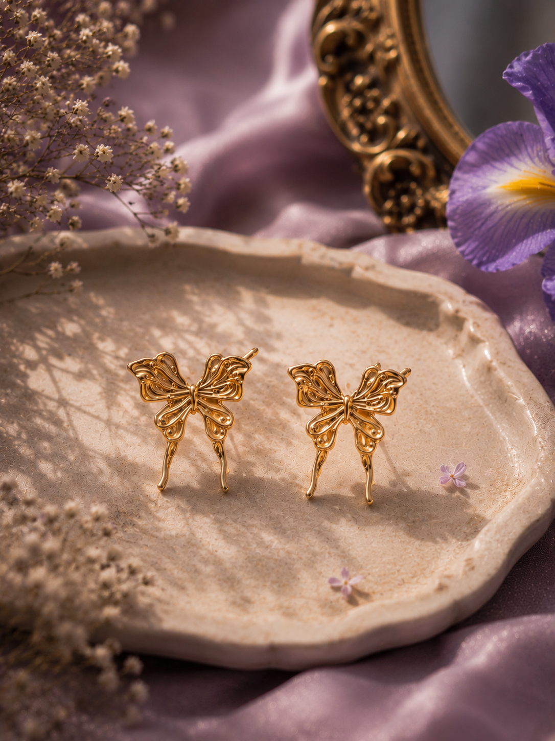 Gold butterfly stud earrings displayed on a ceramic tray with lavender flowers and soft silk fabric styling.
