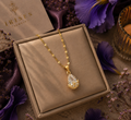 A gold necklace with a teardrop-shaped clear gemstone pendant displayed inside a beige jewelry box, surrounded by purple flowers and a candle.