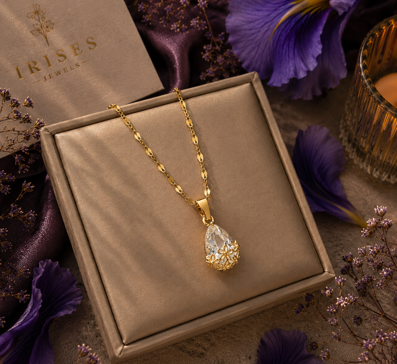 A gold necklace with a teardrop-shaped clear gemstone pendant displayed inside a beige jewelry box, surrounded by purple flowers and a candle.