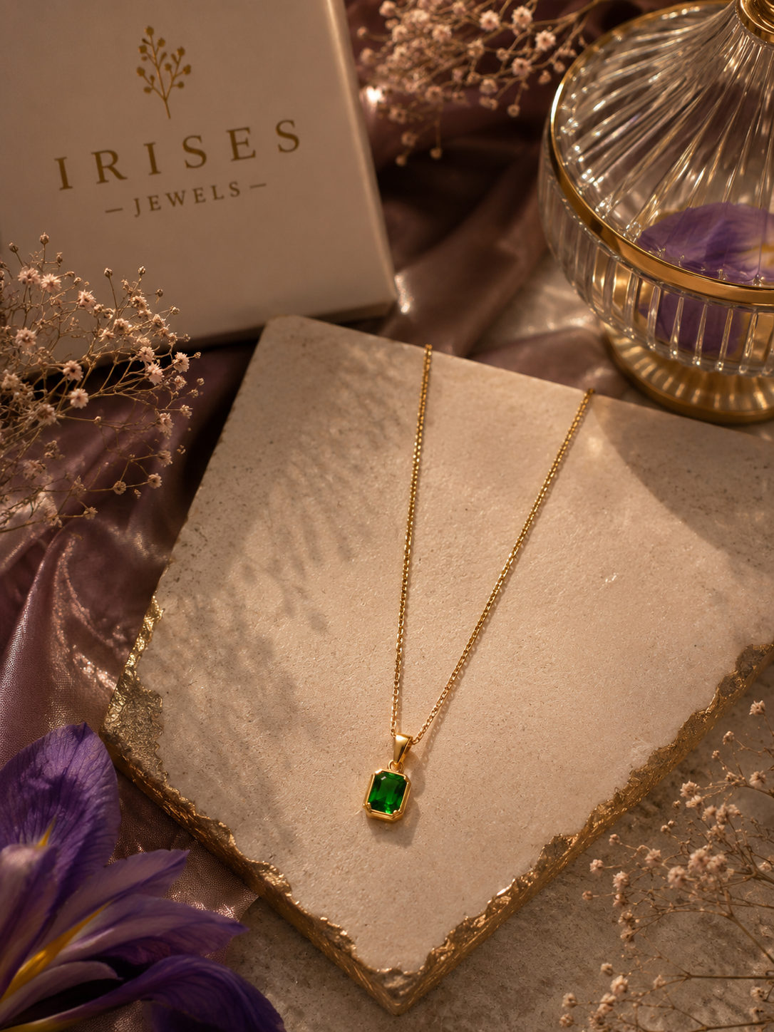 A gold chain necklace with a rectangular green gemstone pendant displayed on a stone slab, with an Irises Jewels box, purple flowers, and a glass candle holder in the background.