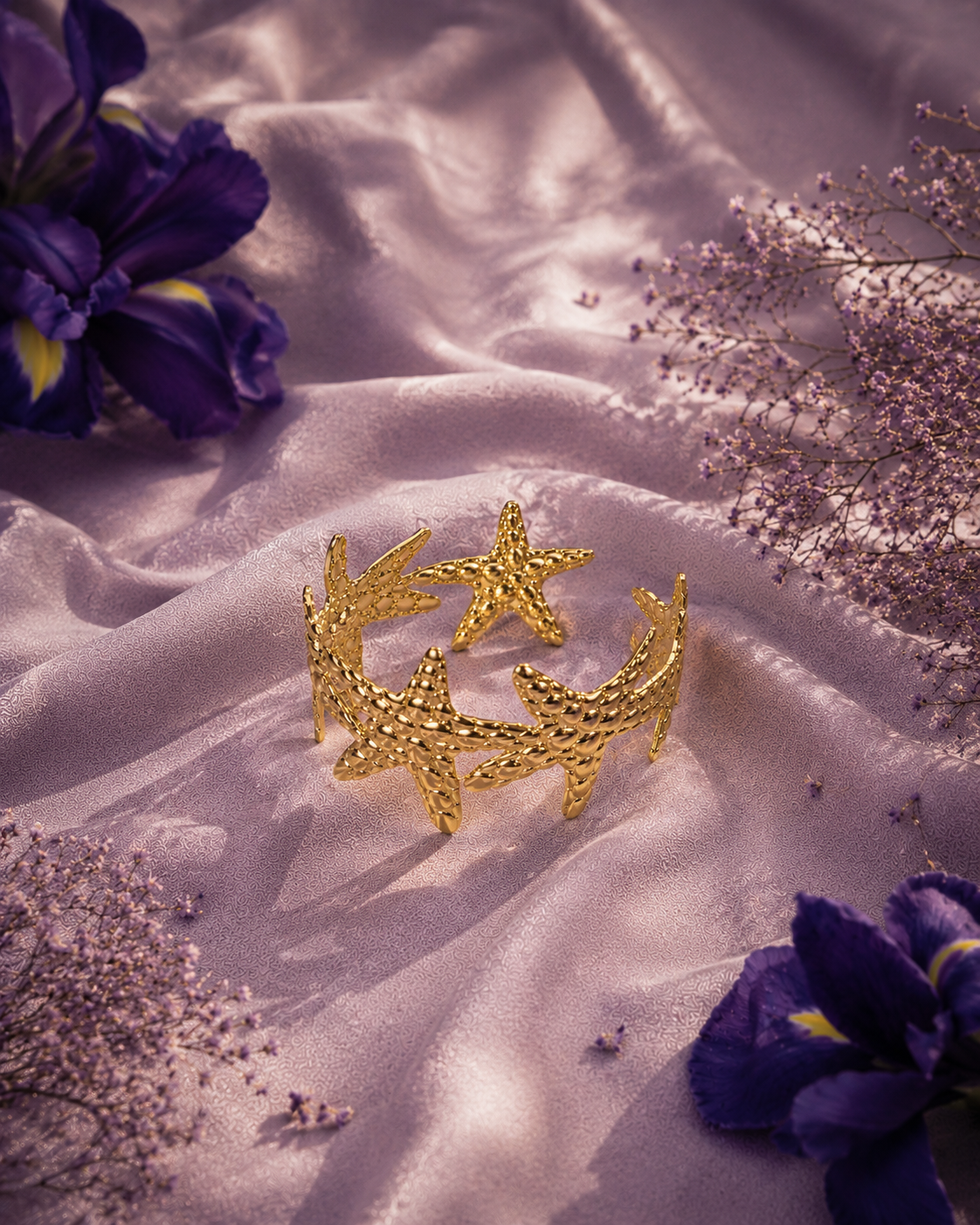 A gold starfish-shaped cuff bracelet displayed on draped lavender satin fabric, surrounded by purple flowers and dried baby's breath.