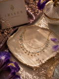 A gold-toned double-strand necklace with small turquoise-blue stones and dangling pearl-like beads, displayed on a metallic shell tray. The brand box 'IRISES JEWELS' is visible in the upper left, with purple flowers and decorative props around it.