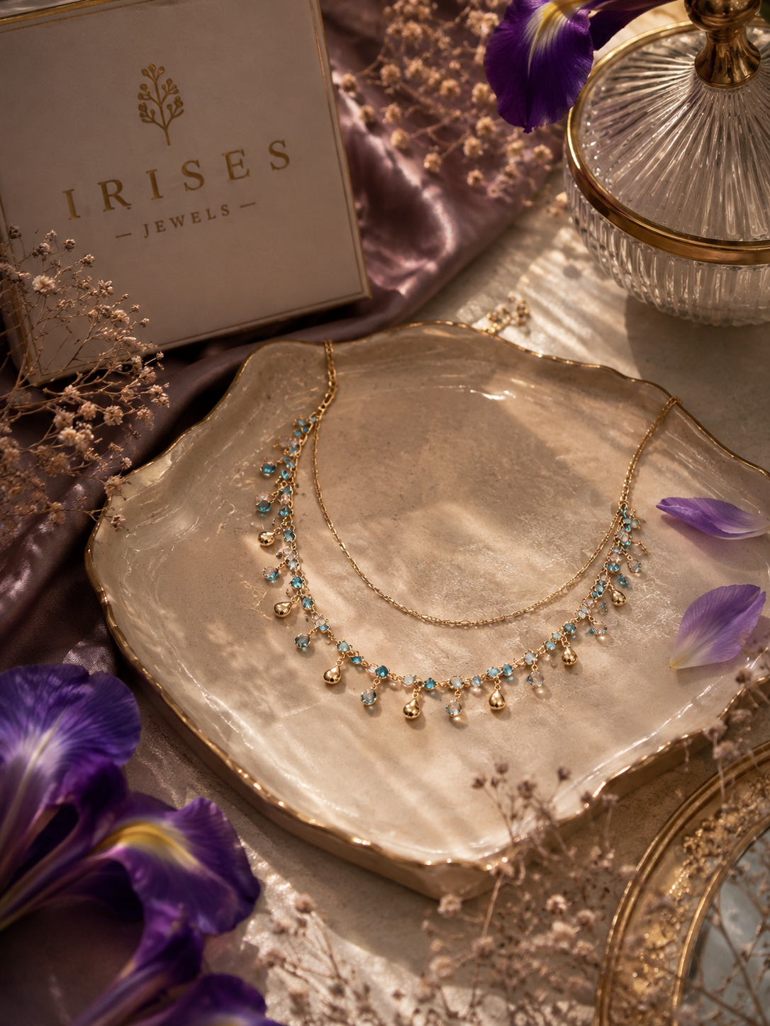 A gold-toned double-strand necklace with small turquoise-blue stones and dangling pearl-like beads, displayed on a metallic shell tray. The brand box 'IRISES JEWELS' is visible in the upper left, with purple flowers and decorative props around it.
